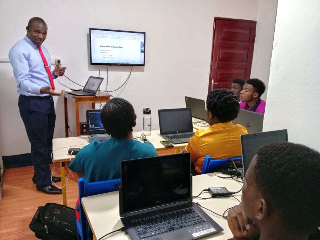 Instructor in blue shirt and red tie teaches students on laptops in a small computer classroom with wall-mounted screen.